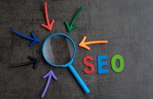 The word "SEO" in red, blue and green on blackboard next to magnifying glass with arrows pointing toward it.